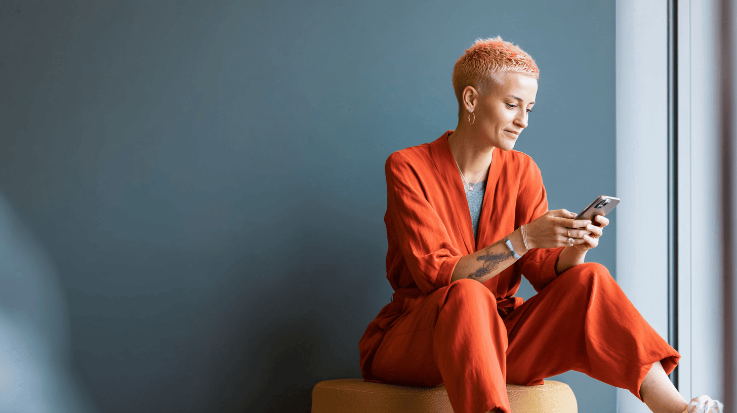A person with short pink hair sits on an ottoman, wearing an orange outfit and using a smartphone against a blue wall.