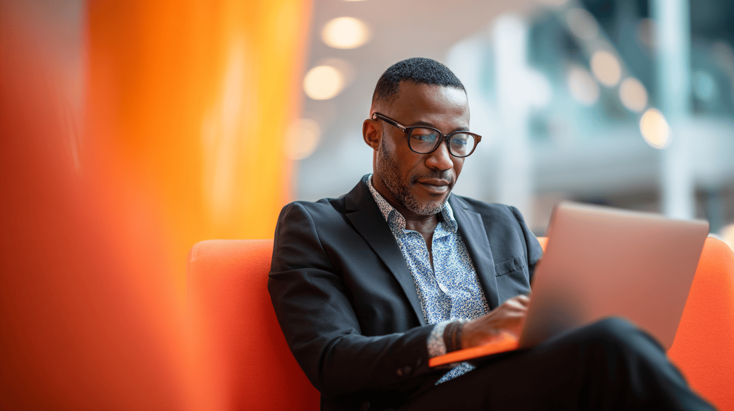 A man in a suit working on a laptop while sitting on an orange couch in a modern, well-lit space.