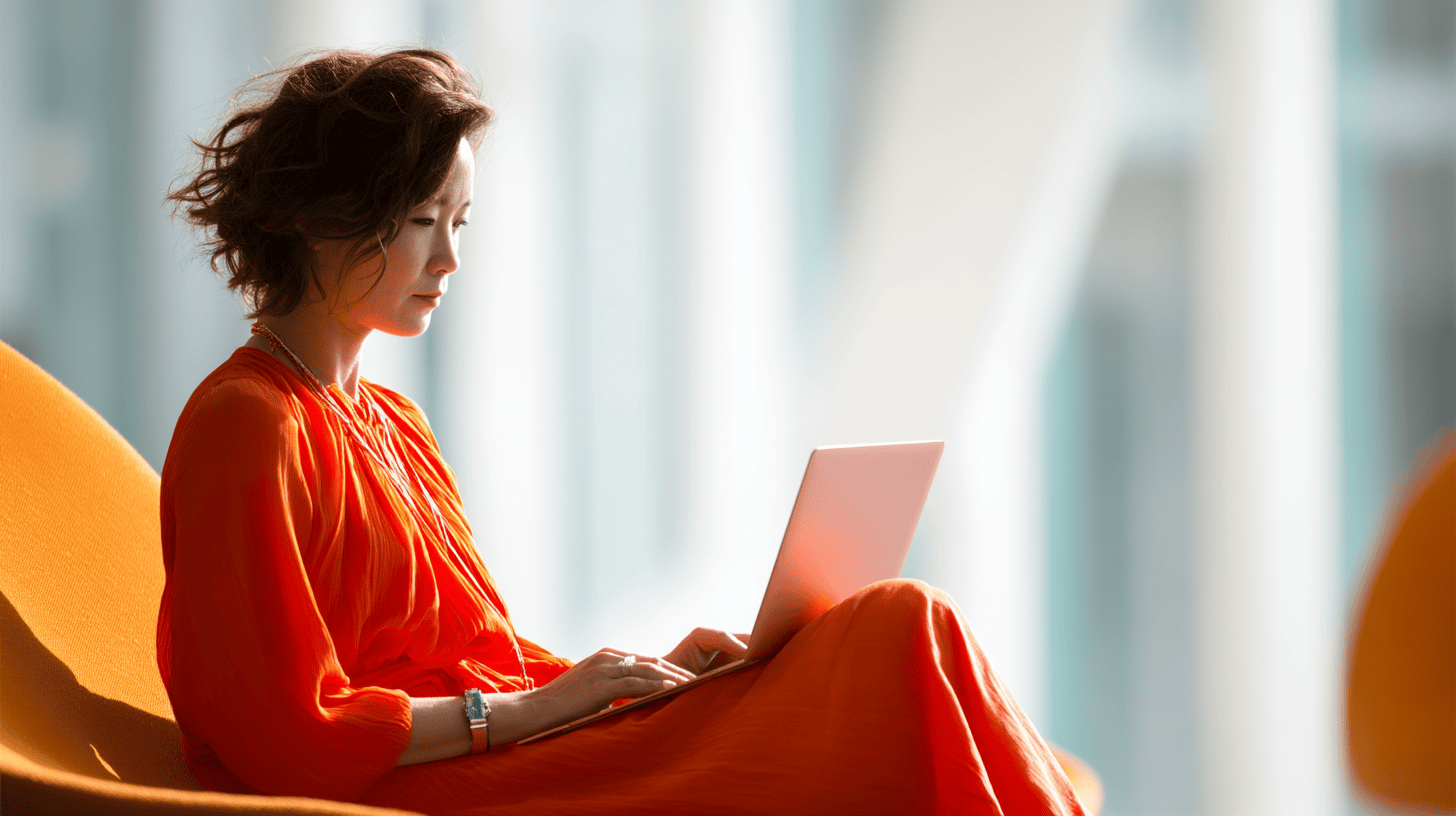 A woman in an orange dress sits on a chair, focused on her laptop in a bright, modern environment.