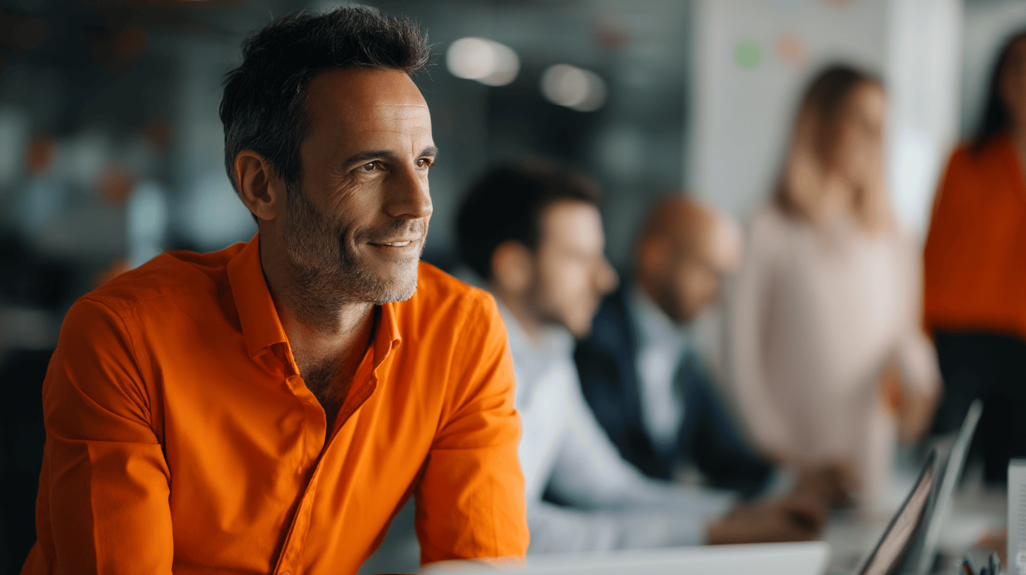 A man in an orange shirt smiles, seated at a table with blurred colleagues working on laptops in an office setting.