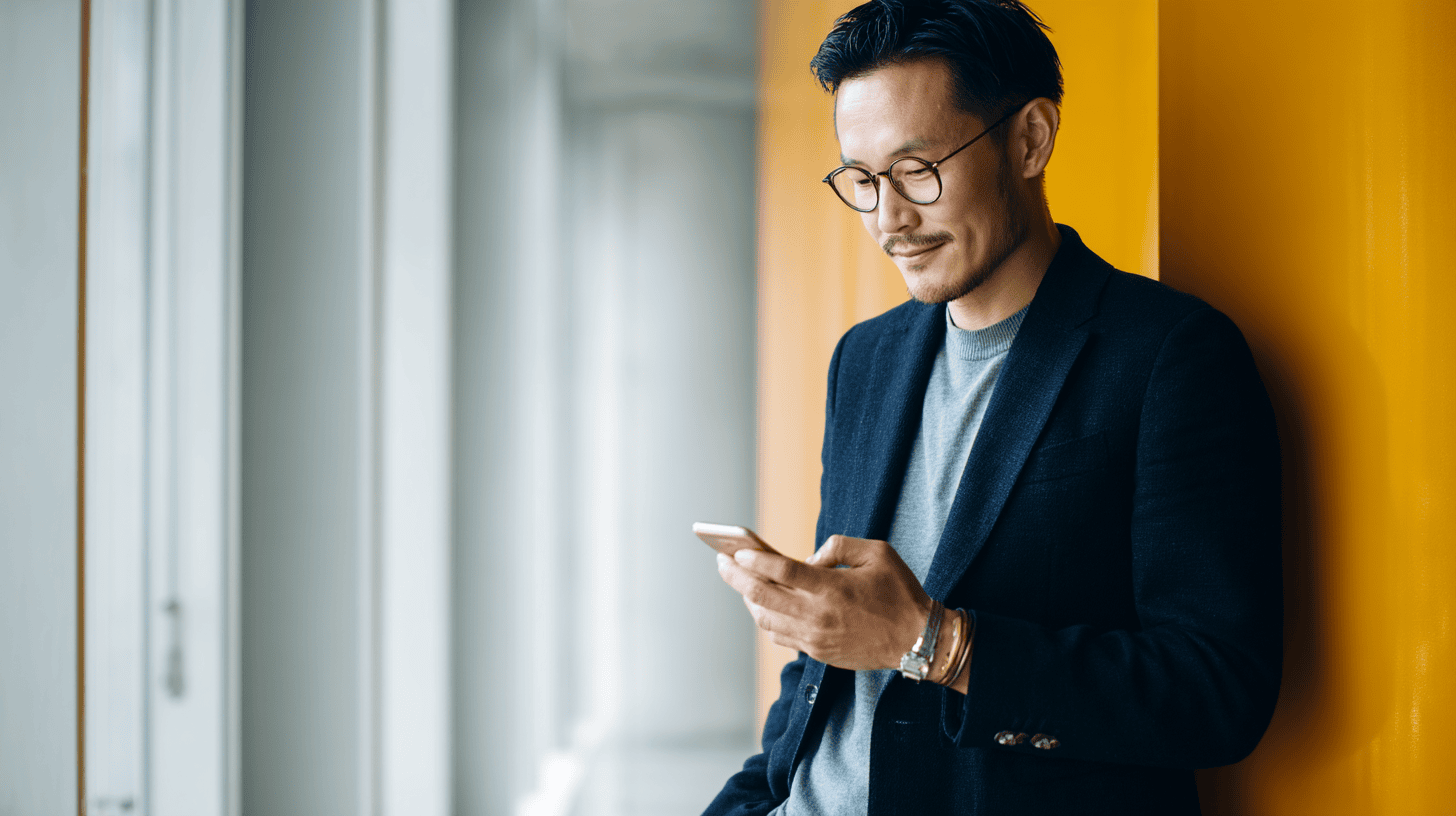 A man in a blazer checks his phone while leaning against a bright yellow wall in a modern interior.