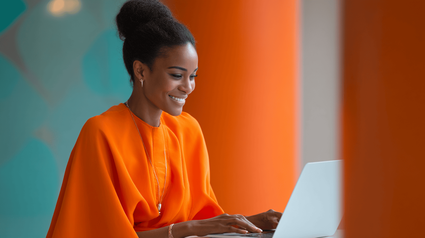 A woman in an orange top works on a laptop with a bright background, smiling as she types.