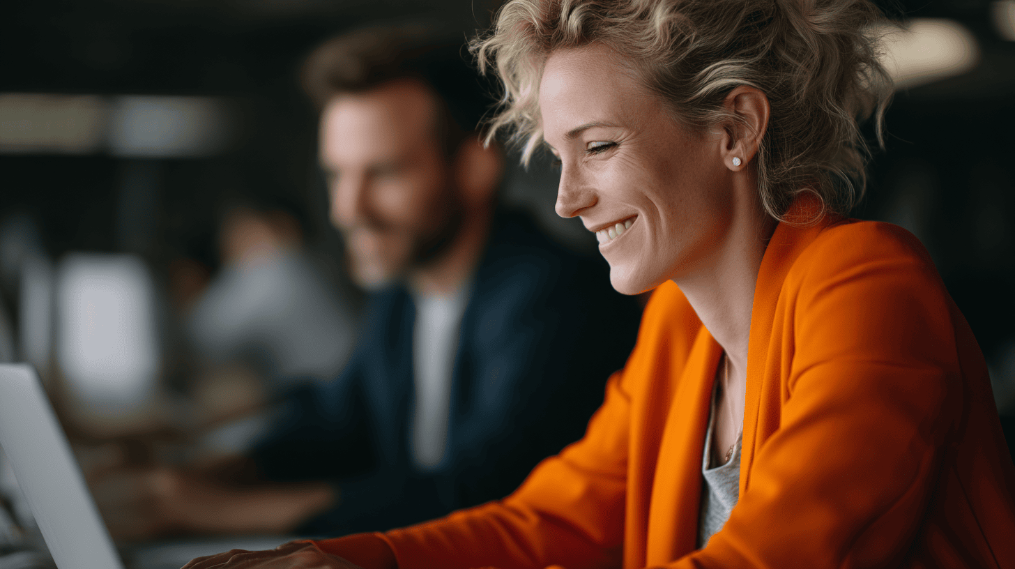 A woman in a bright orange blazer smiles while working on a laptop; a blurred man is seated nearby.