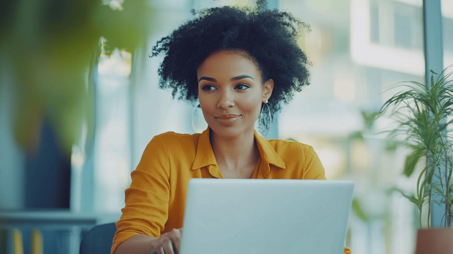 A woman with curly hair sits at a desk, smiling while looking at a laptop, surrounded by plants in a bright environment.