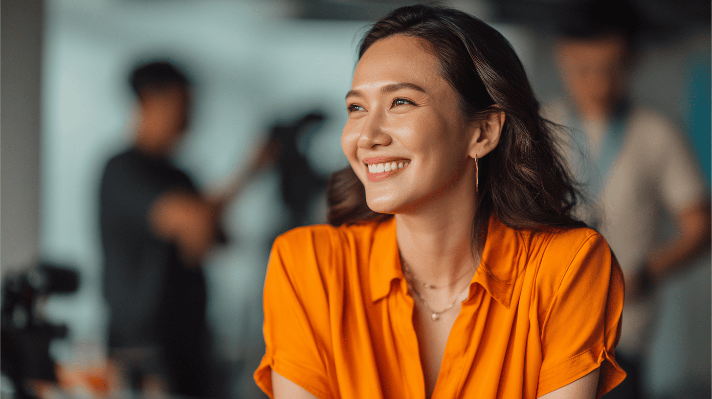 A woman in an orange shirt smiles, with a blurred background of people and camera equipment.