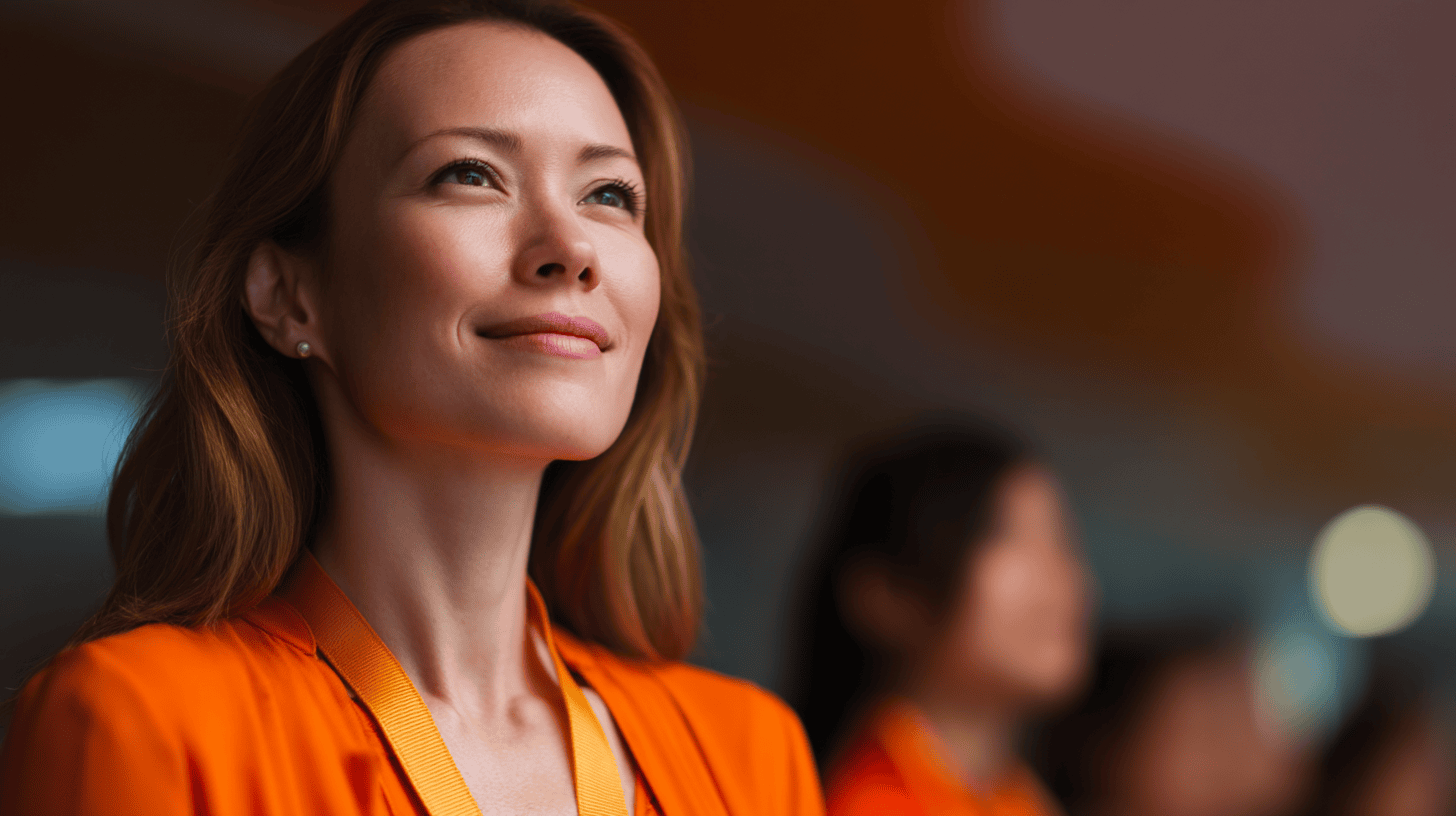 A woman in an orange outfit smiles confidently, with a blurred audience in the background and soft lighting.