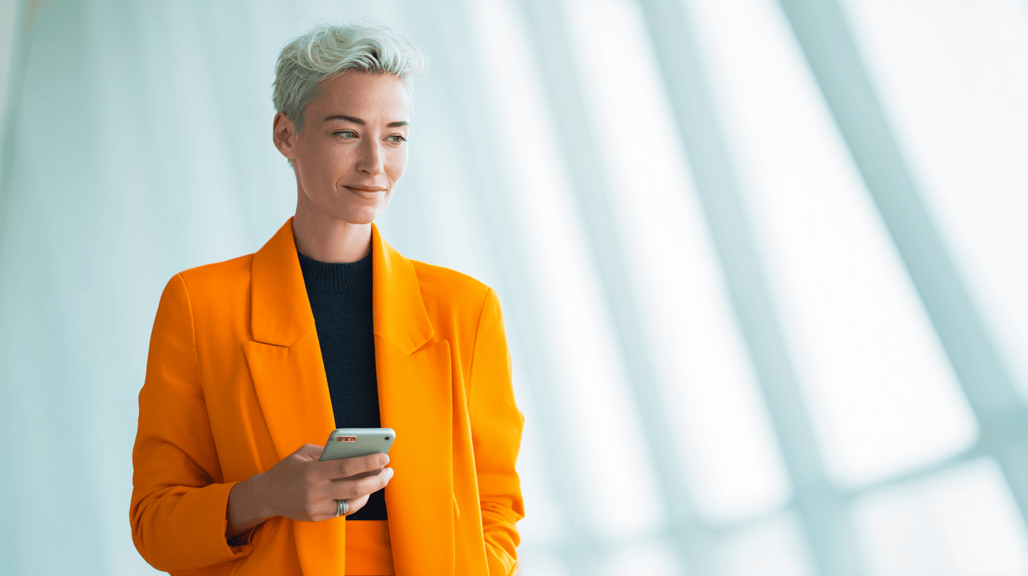 A woman in an orange blazer holds a smartphone, smiling against a backdrop of light-colored geometric windows.