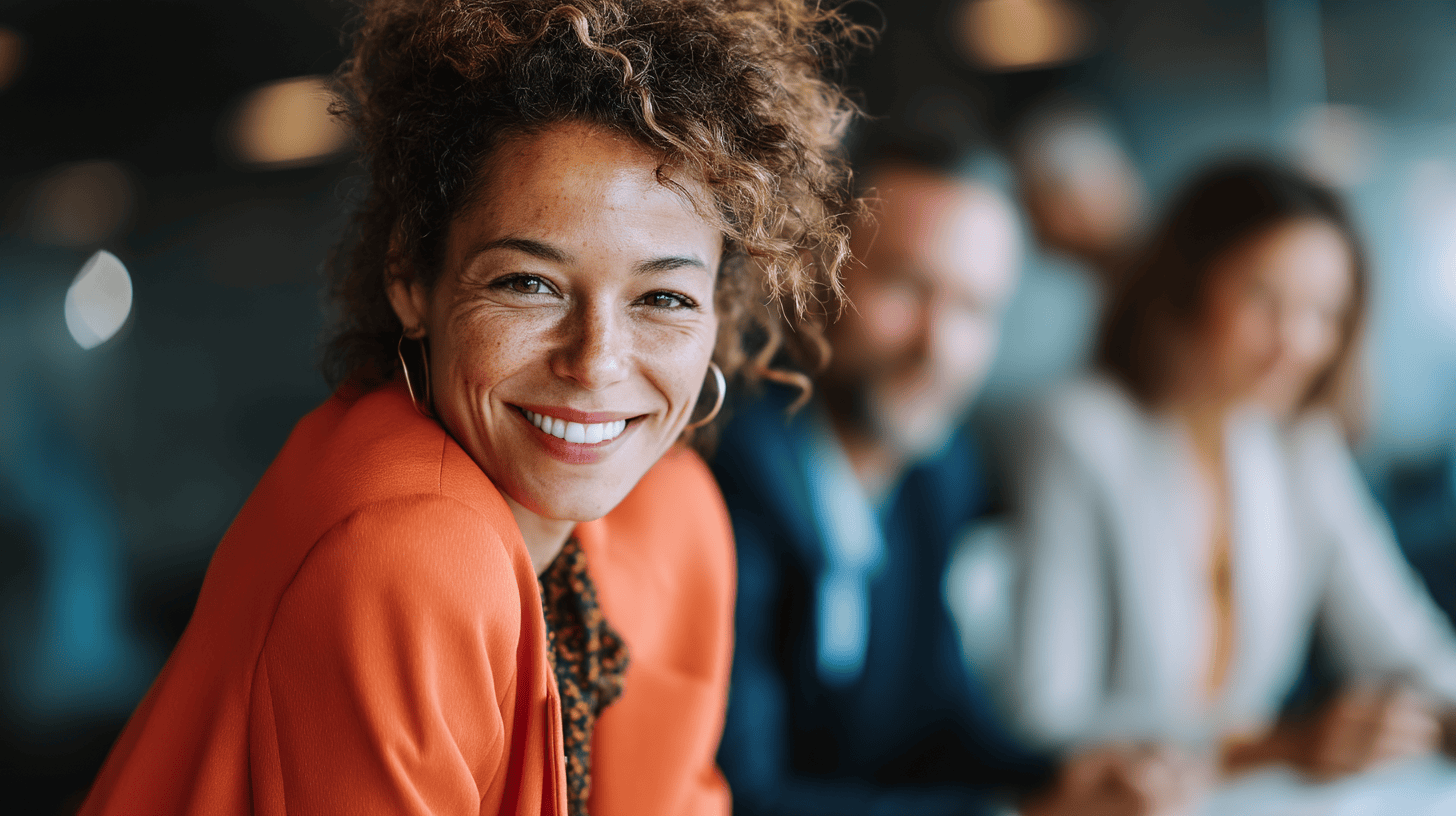 Smiling woman in an orange blazer sits at a conference table with blurred colleagues in the background.