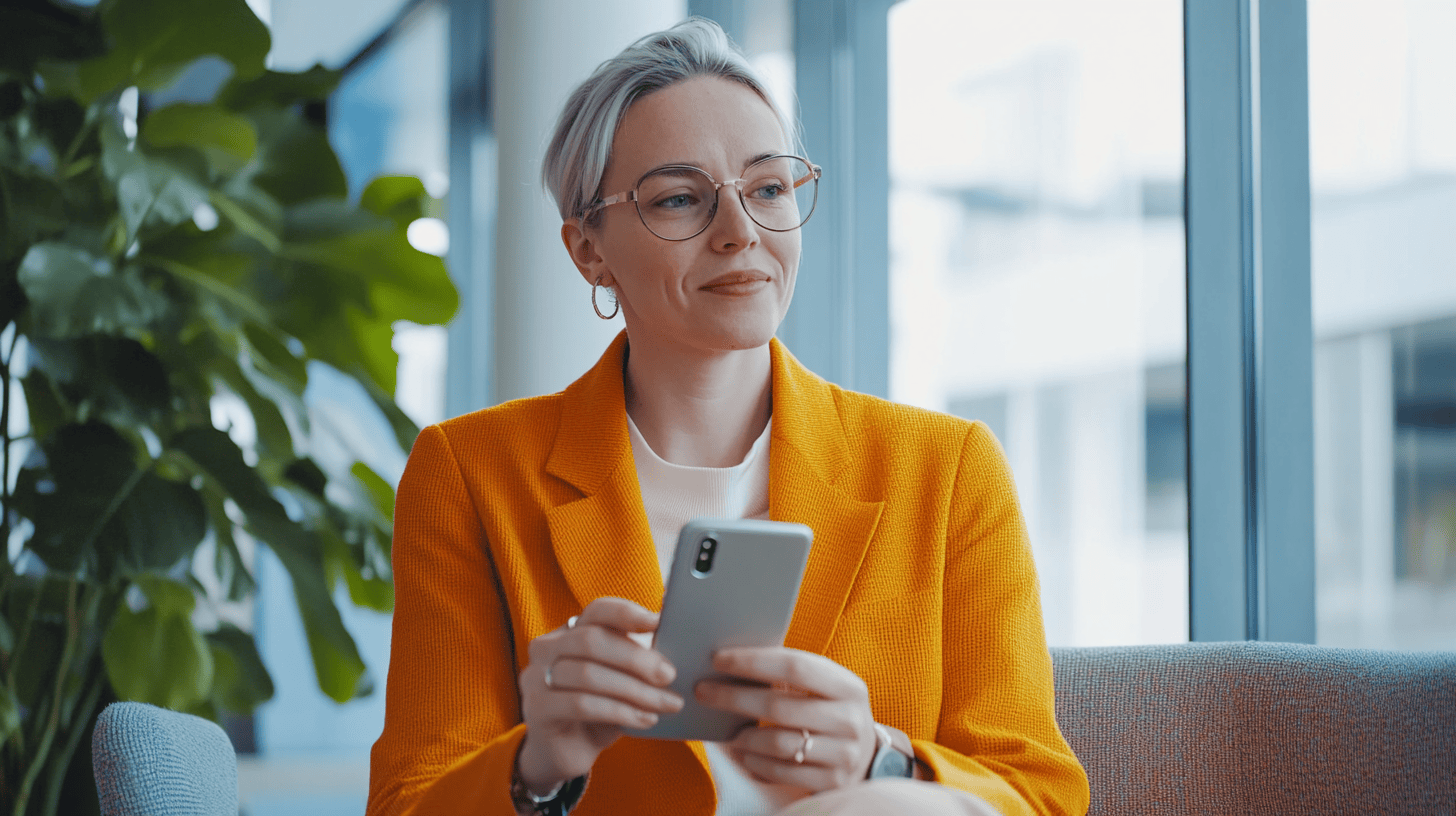 A woman with short, blonde hair and glasses sits on a couch, holding a smartphone, with plants and large windows in the background.