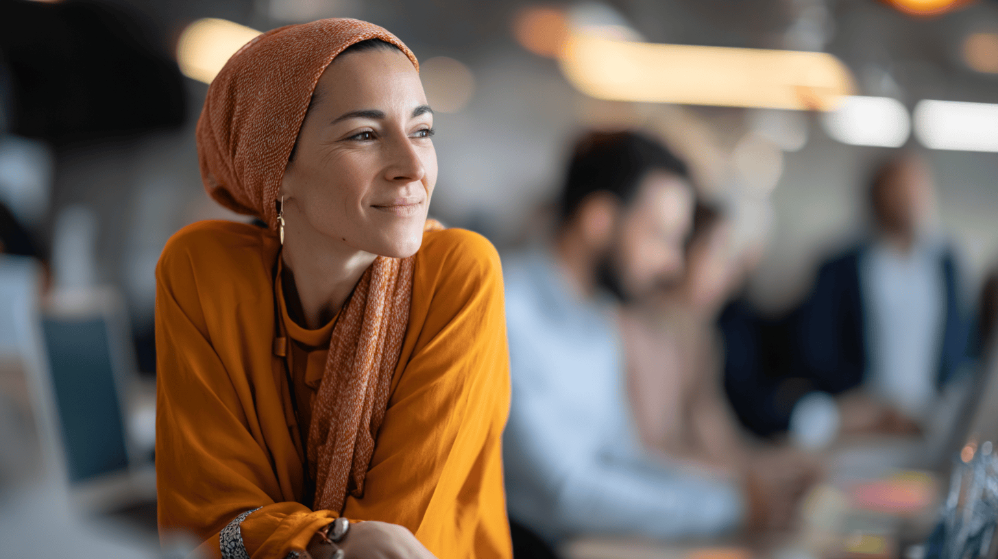 A woman in an orange blouse and patterned headscarf smiles thoughtfully at a busy modern workspace.
