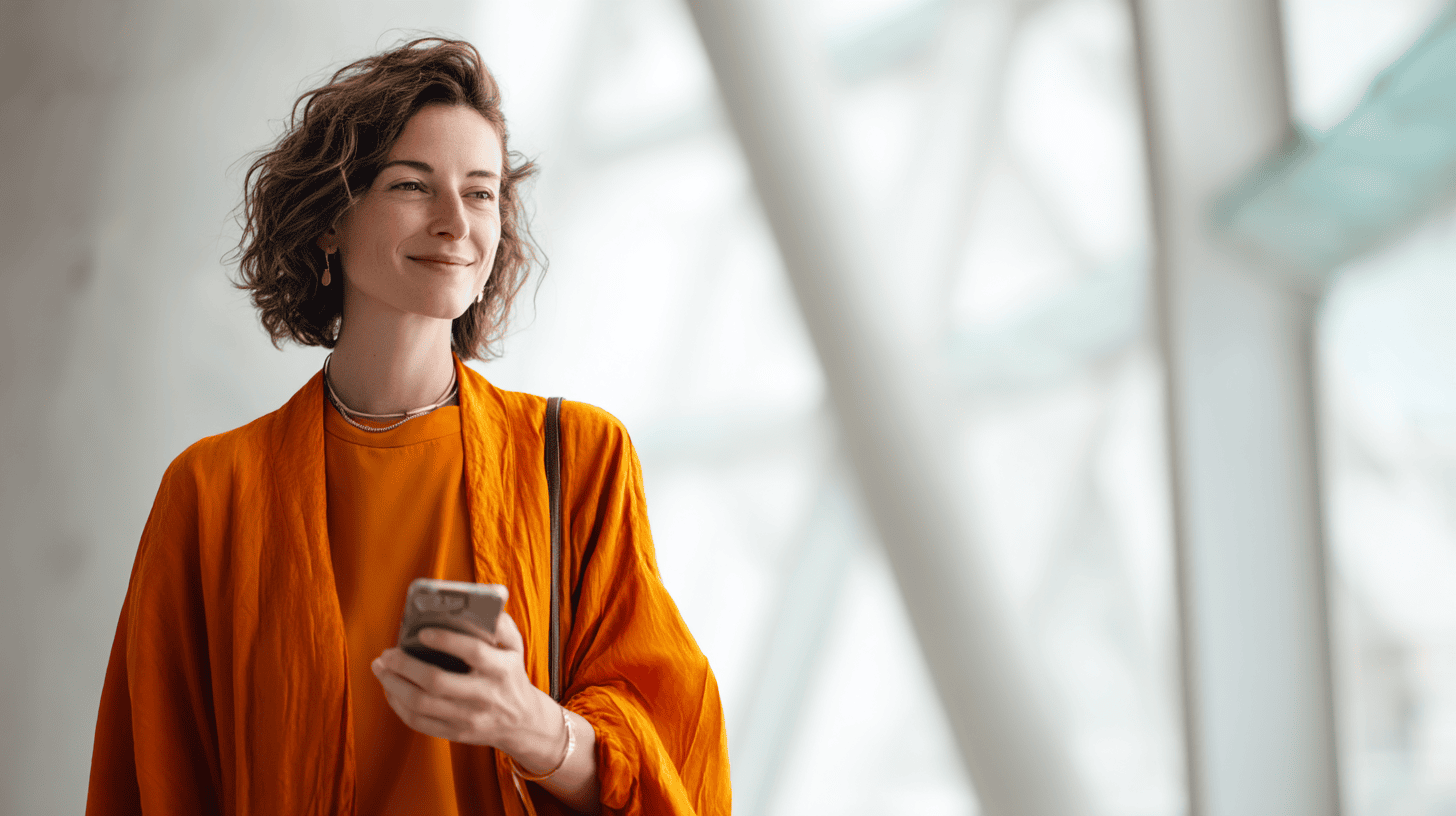 A woman with curly hair is smiling while holding a smartphone, wearing an orange outfit in a modern, bright setting.