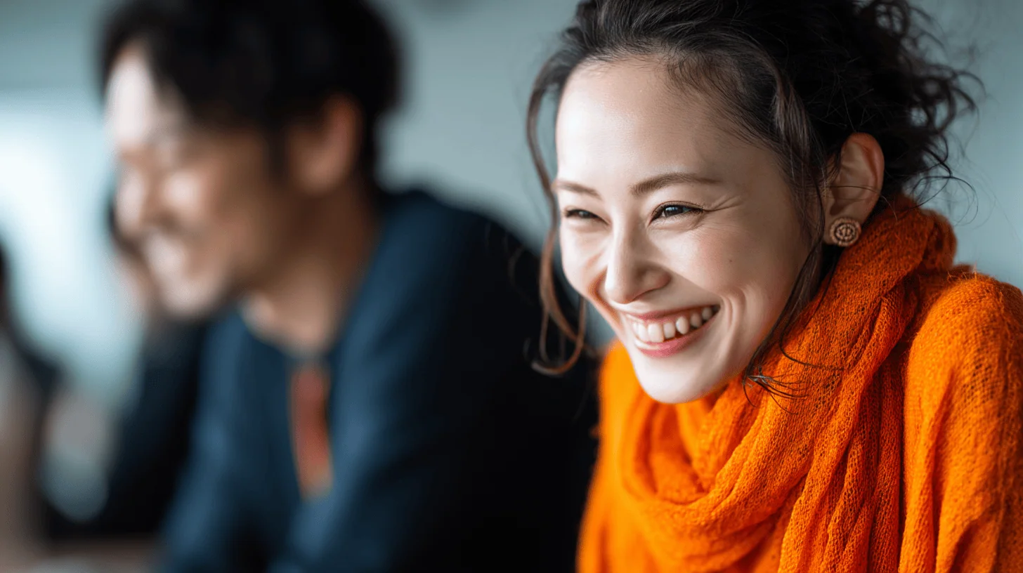 A woman in an orange scarf smiles brightly, with a blurred figure beside her, set in a casual indoor environment.