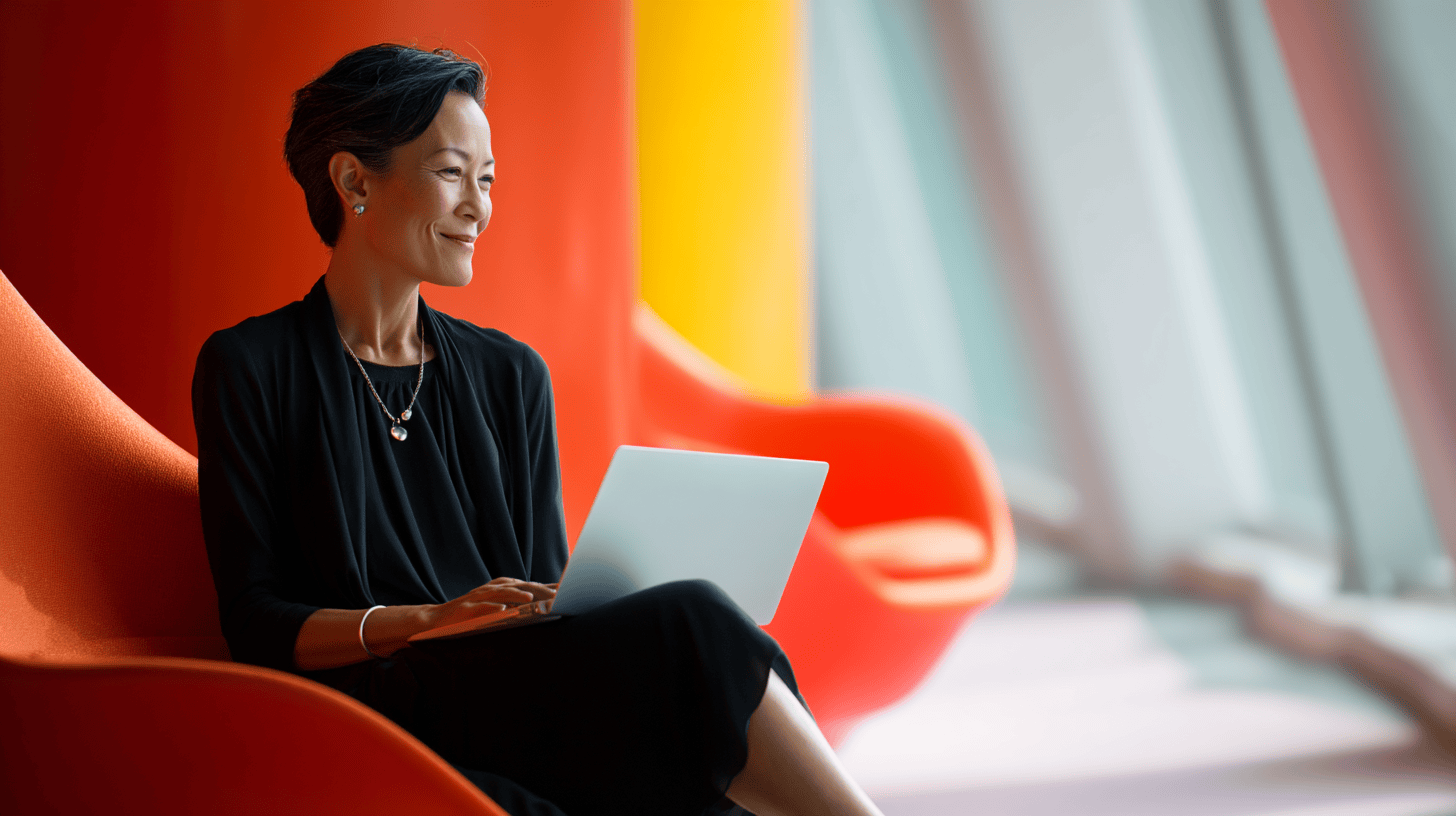 A woman sits in a bright orange chair, using a laptop, with a vibrant backdrop of colorful walls and natural light.