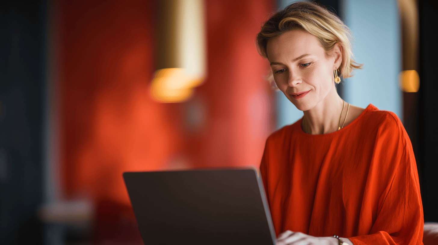 A woman in an orange top works on a laptop in a stylish, modern space with warm, blurred backgrounds.