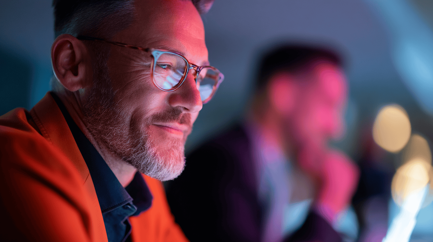 A close-up of a man in an orange jacket, smiling with glasses, in a colorful, softly lit environment.