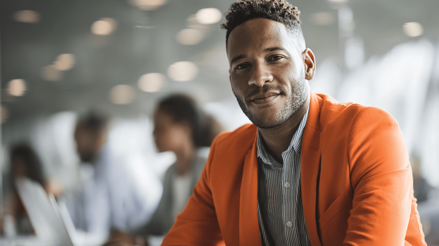 A man in an orange blazer smiles, seated at a table with blurred colleagues in a modern office setting.