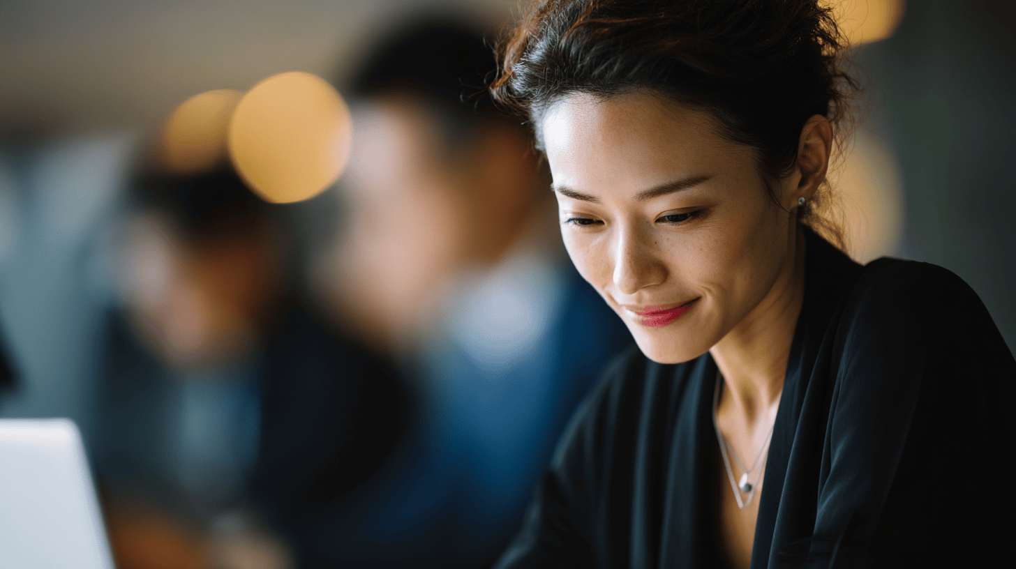 A woman in a black top smiles while using a laptop, with blurred individuals and soft background lights visible.