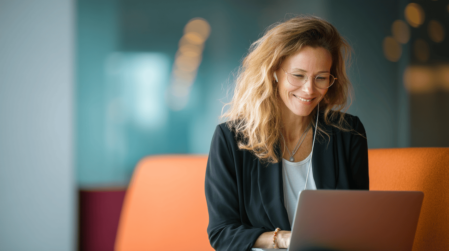 Smiling woman with curly hair, wearing glasses and headphones, sitting on an orange couch, using a laptop.