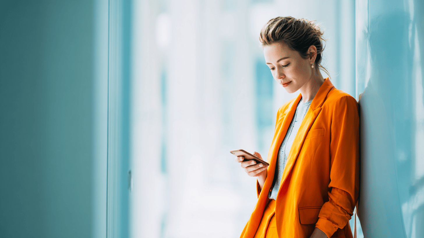 A woman in an orange blazer leans against a light blue wall, looking down at her smartphone in a bright, modern space.