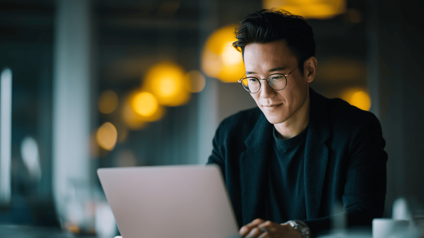 A man with glasses sits at a table, smiling while working on a laptop in a softly lit café.