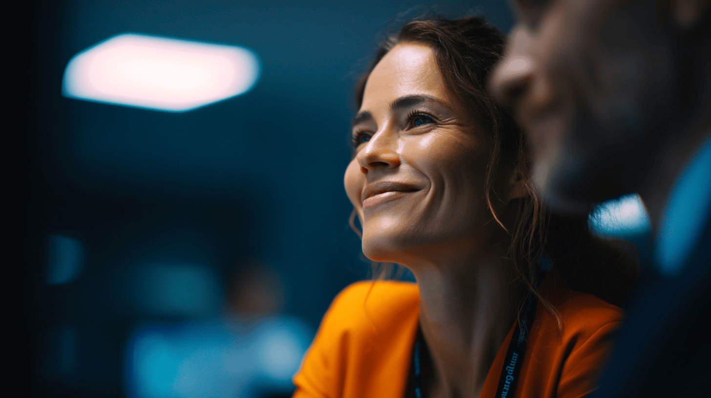 A smiling woman in an orange blazer sits in a dimly lit office, with a blurred colleague beside her.
