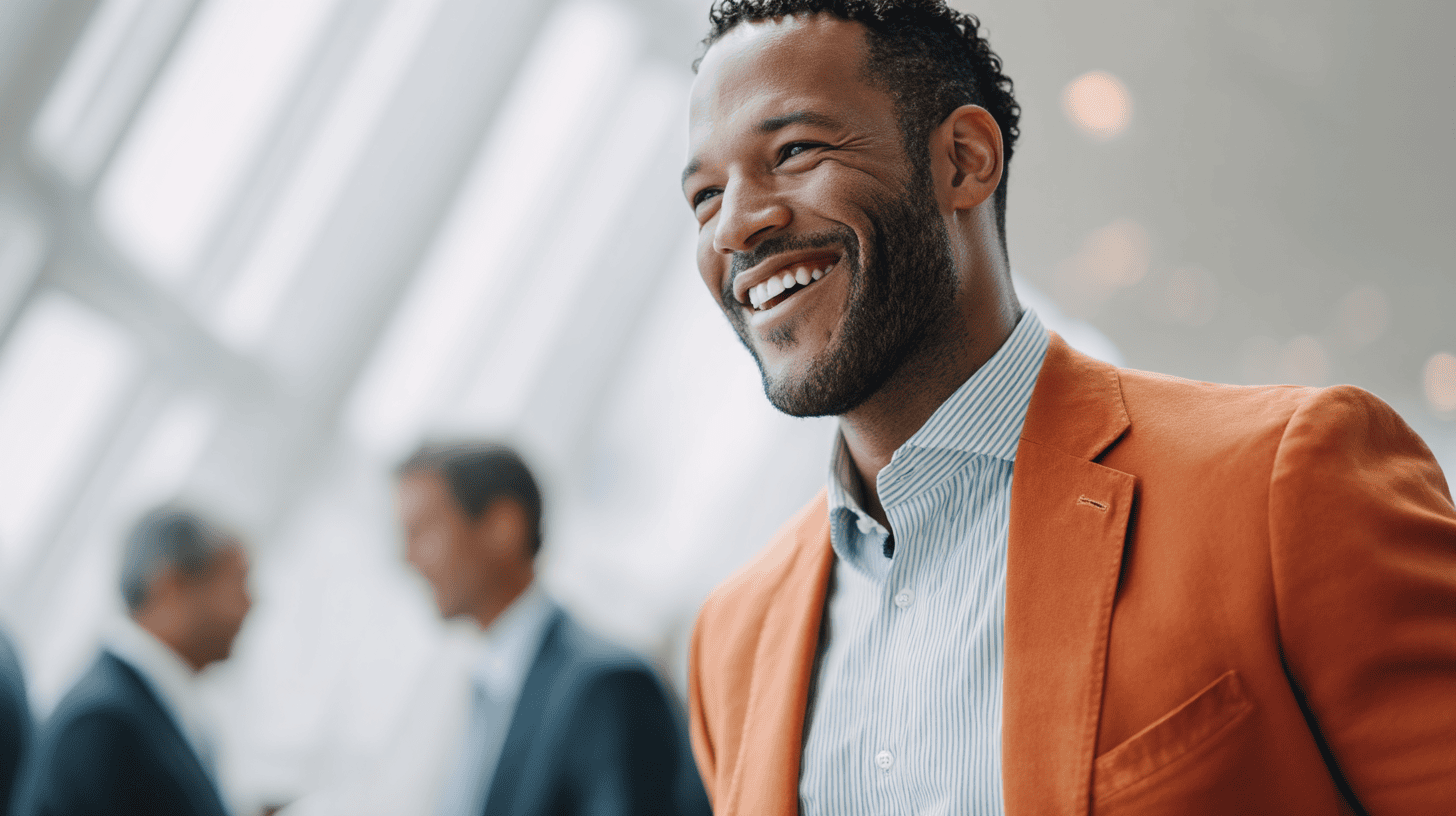 A smiling man in an orange blazer and striped shirt stands in a bright, airy space with blurred figures in the background.