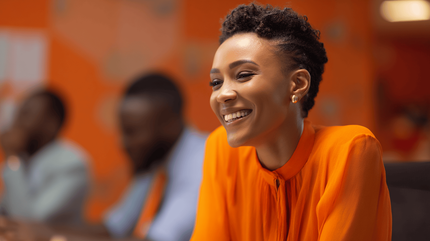 A smiling woman in an orange blouse sits at a meeting table, with blurred colleagues in the background.