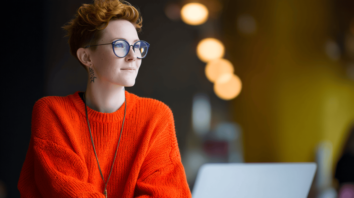 A young person with short, curly hair wearing glasses and an orange sweater, looking thoughtfully out of a window.