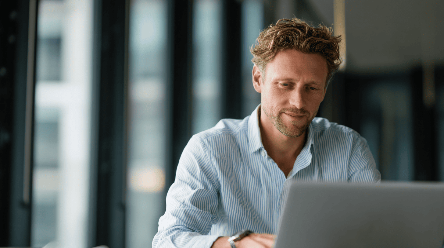 A man with curly hair sits at a desk, focused on a laptop, with large windows and blurred cityscape in the background.