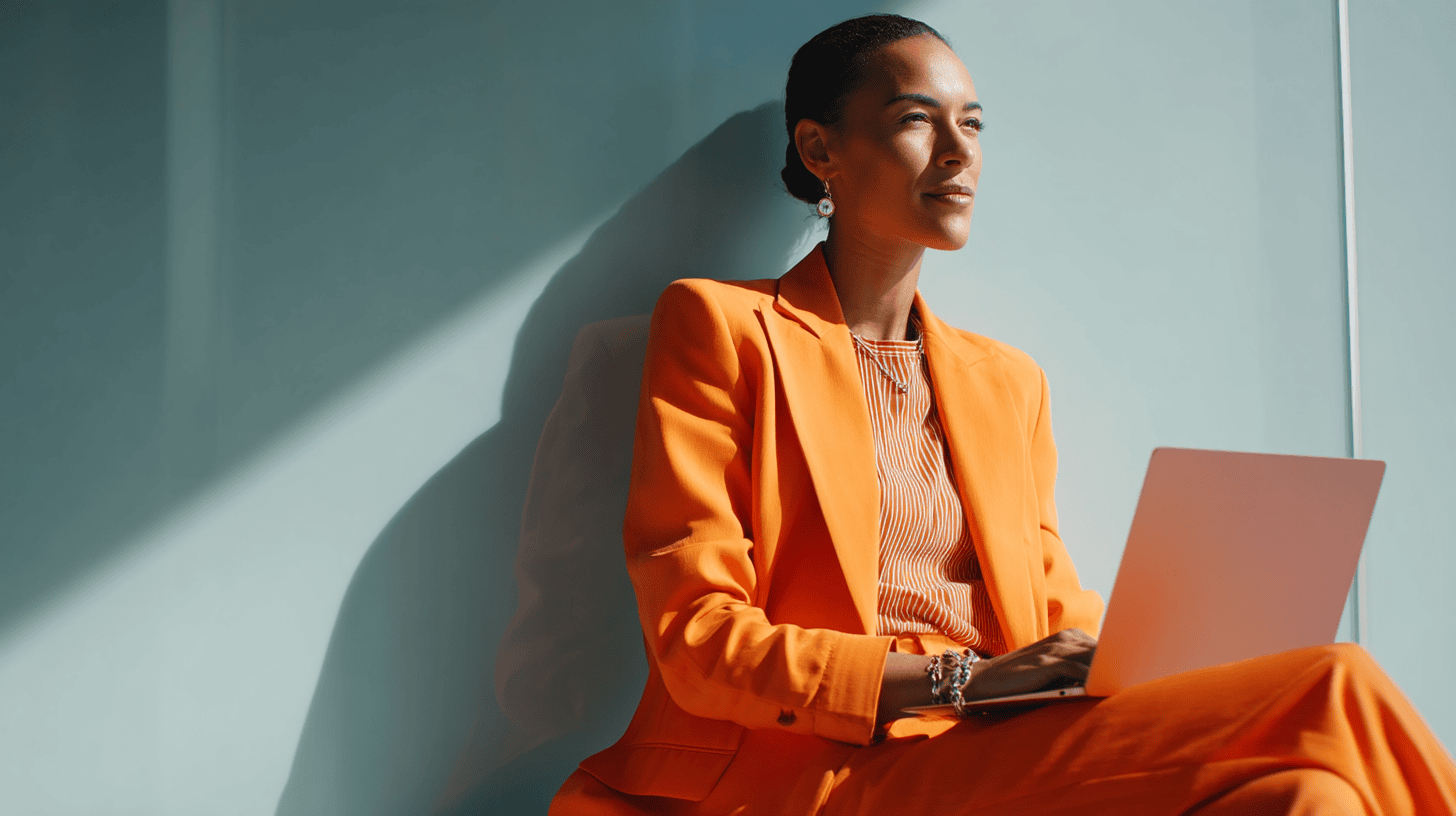 A woman in an orange suit sitting against a wall, looking thoughtfully at a laptop. Sunlight casts soft shadows.