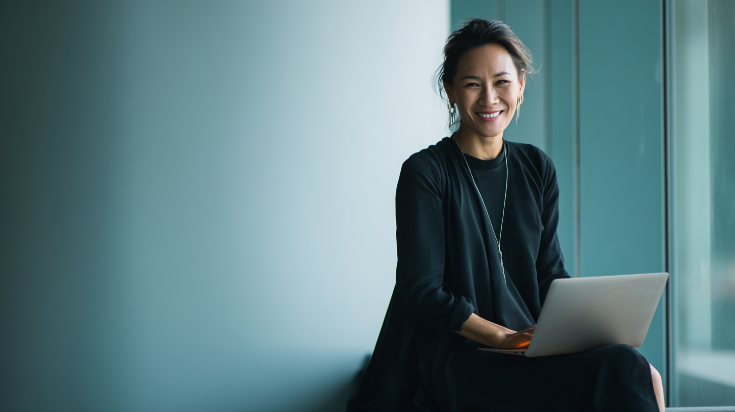 A woman in a black outfit sits with a laptop, smiling, against a soft blue background by a window.