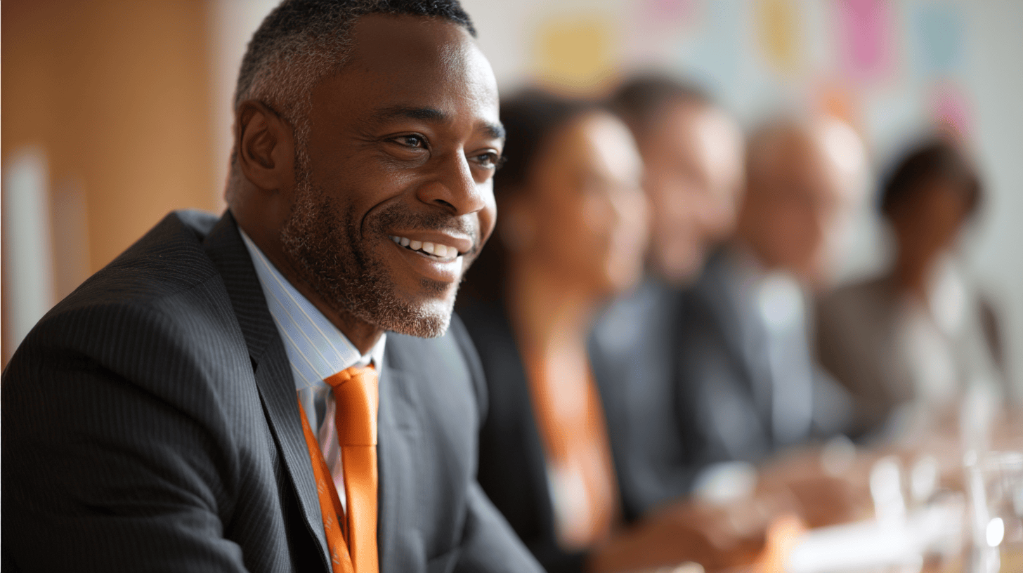 A smiling man in a suit with an orange tie sits at a conference table, with blurred colleagues in the background.