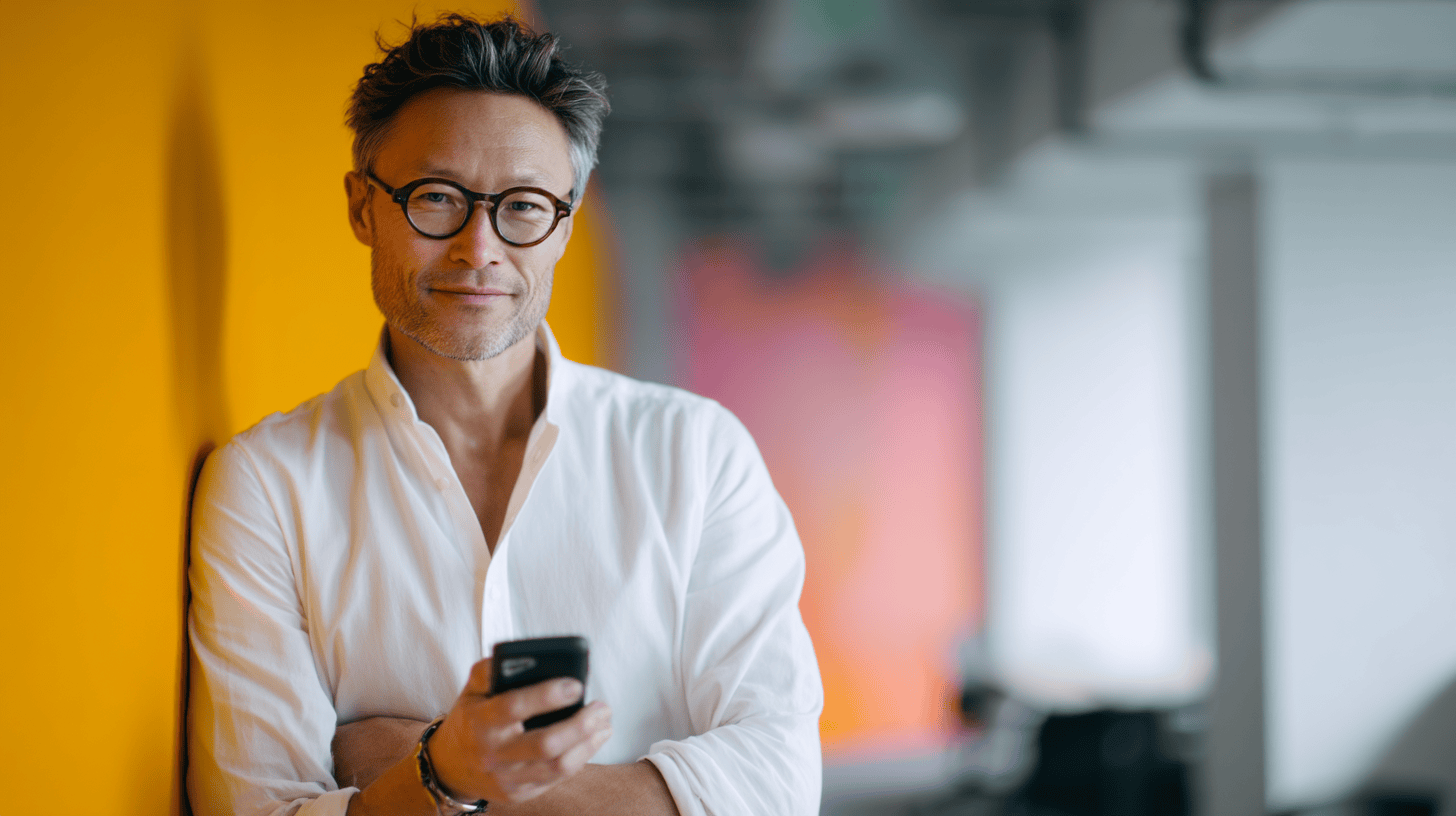 A man with glasses wears a white shirt and smiles while holding a smartphone against a bright orange background.
