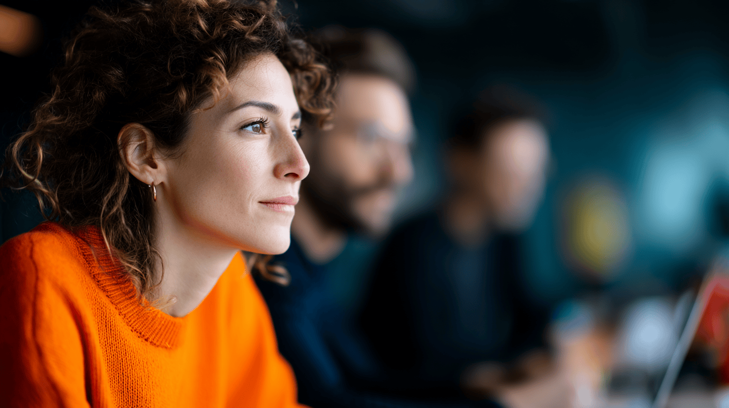 A woman with curly hair in an orange sweater sits thoughtfully, with two blurred colleagues in the background.