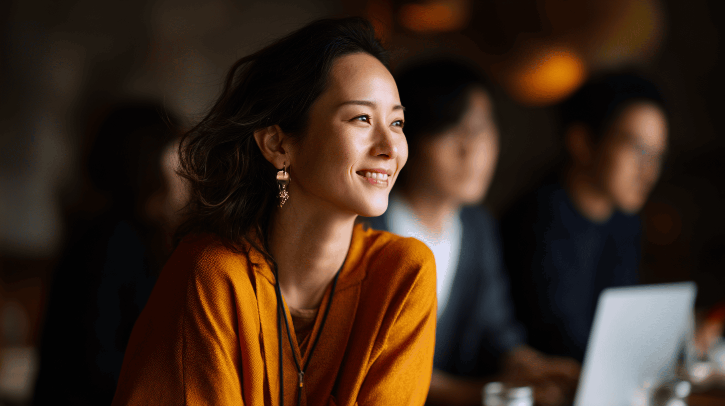 A woman smiles while seated at a cafe, wearing an orange sweater and earrings, with blurred figures in the background.