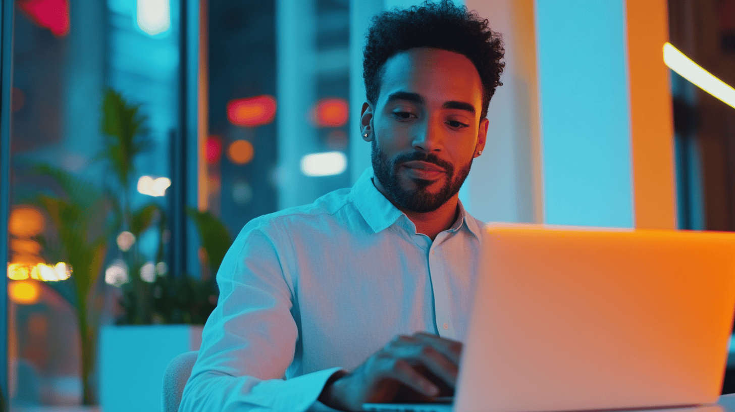 A man with curly hair types on a laptop in a brightly lit, modern workspace with colorful ambient lighting.