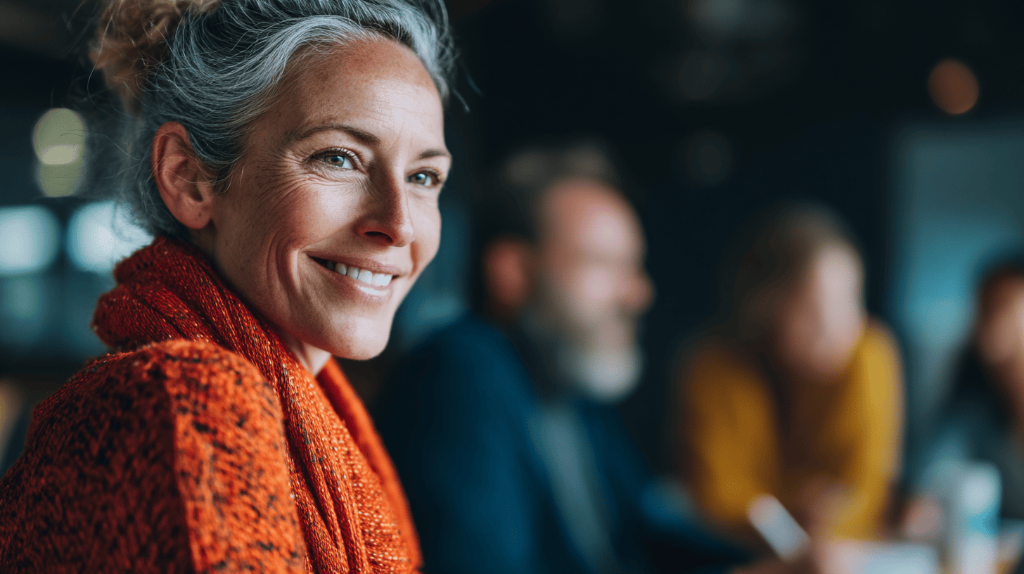 A woman with gray hair smiles warmly, wearing an orange sweater, with blurred figures in the background.