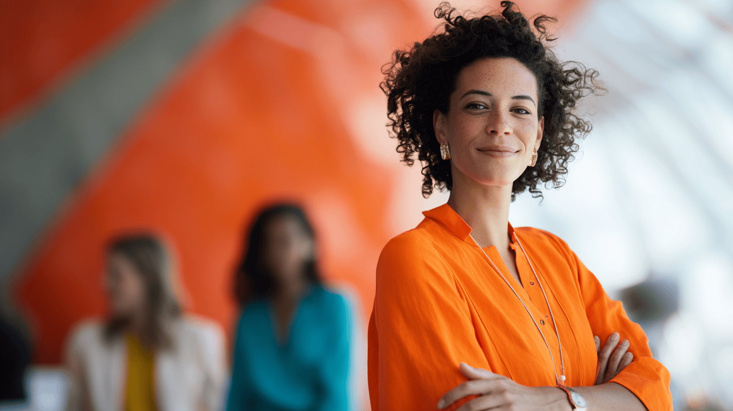 A woman with curly hair smiles confidently, wearing an orange blouse, with an orange and gray background. Two others are blurred behind her.