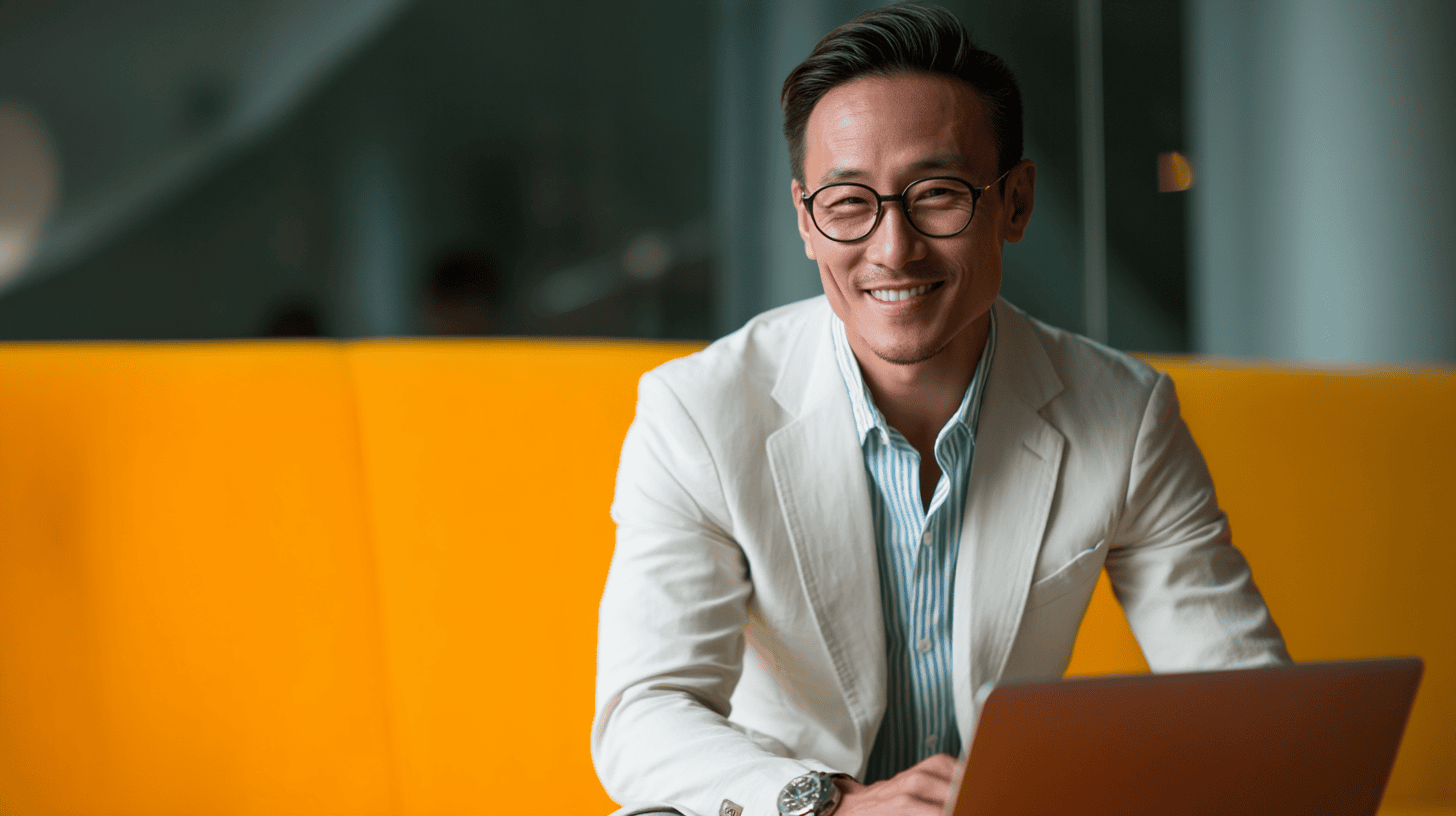 Smiling man in a light blazer and glasses sits at a laptop, with a bright orange background.