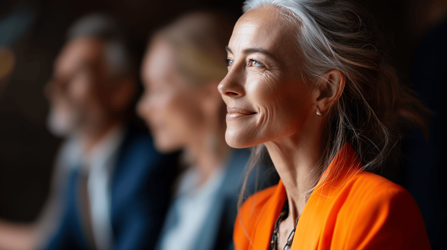 A woman with silver hair smiles thoughtfully while seated; two blurred figures are visible in the background.