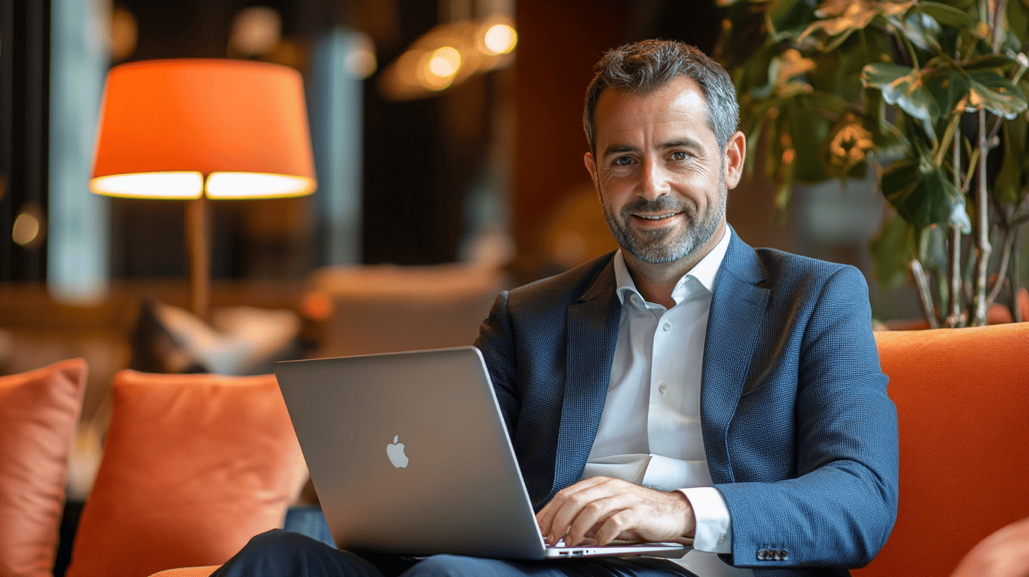 A man with a beard is sitting on an orange sofa, working on a laptop with a lamp and plants in the background.