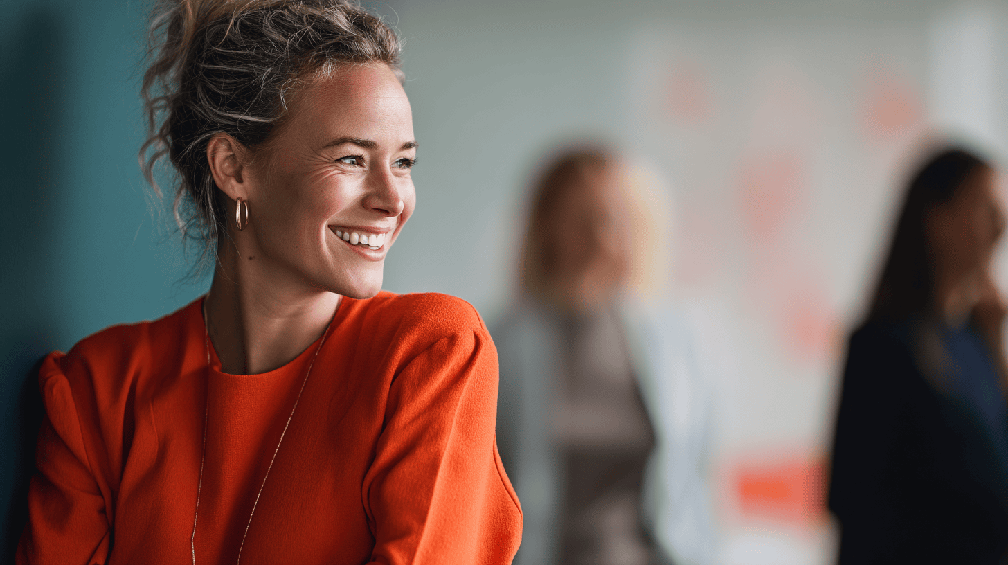 A smiling woman in an orange top stands in the foreground, with blurred figures of other women in the background.