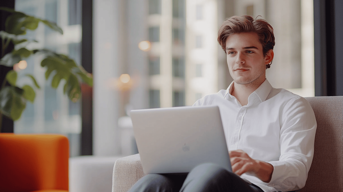 A young man in a white shirt works on a laptop while sitting on a light-colored couch beside a large window.