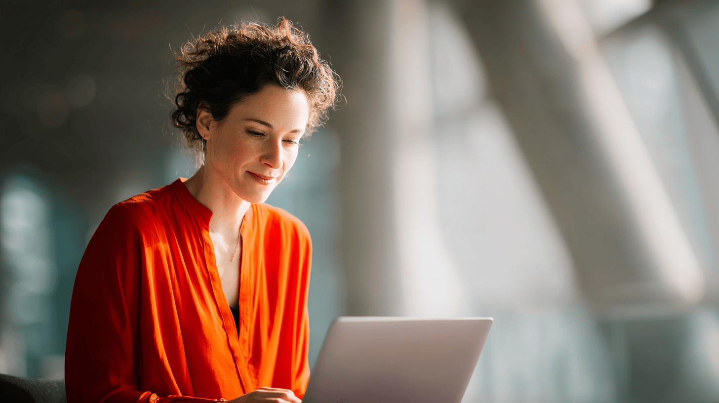 A woman in an orange blouse is smiling while using a laptop in a bright, modern setting.