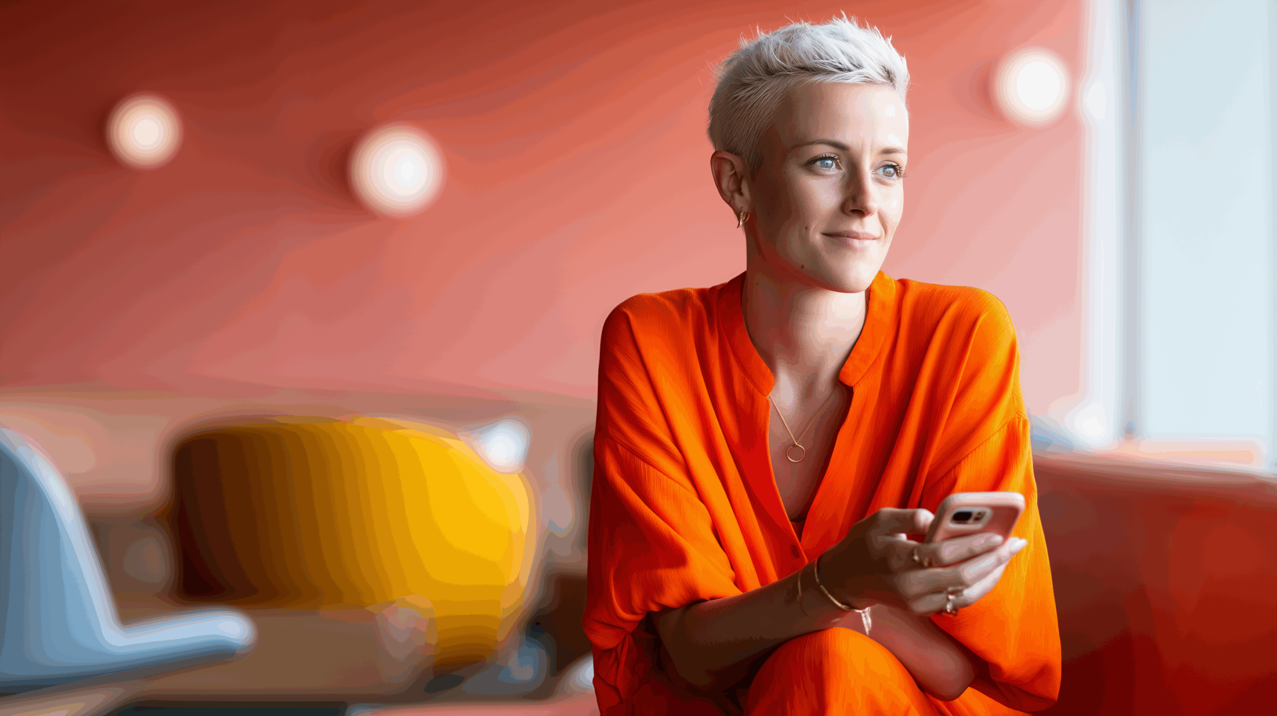 A young woman in an orange outfit sits casually, holding a phone, with a colorful, modern background.