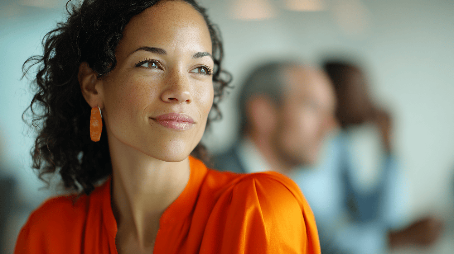 A woman with curly hair, wearing an orange shirt and earrings, smiles softly in a blurred office setting.