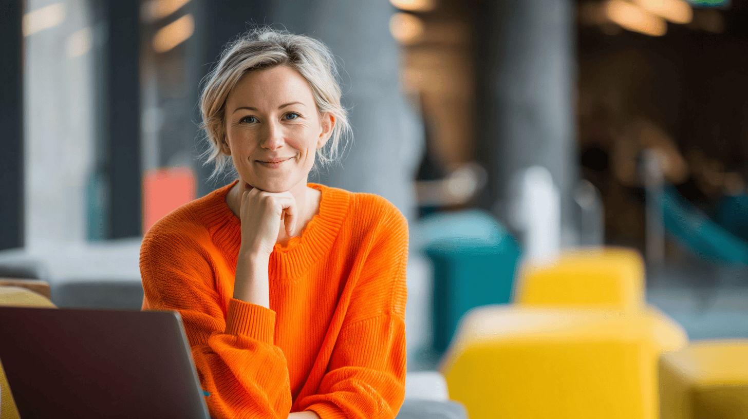 A smiling woman in an orange sweater sits by a laptop, with colorful furniture and a modern workspace in the background.