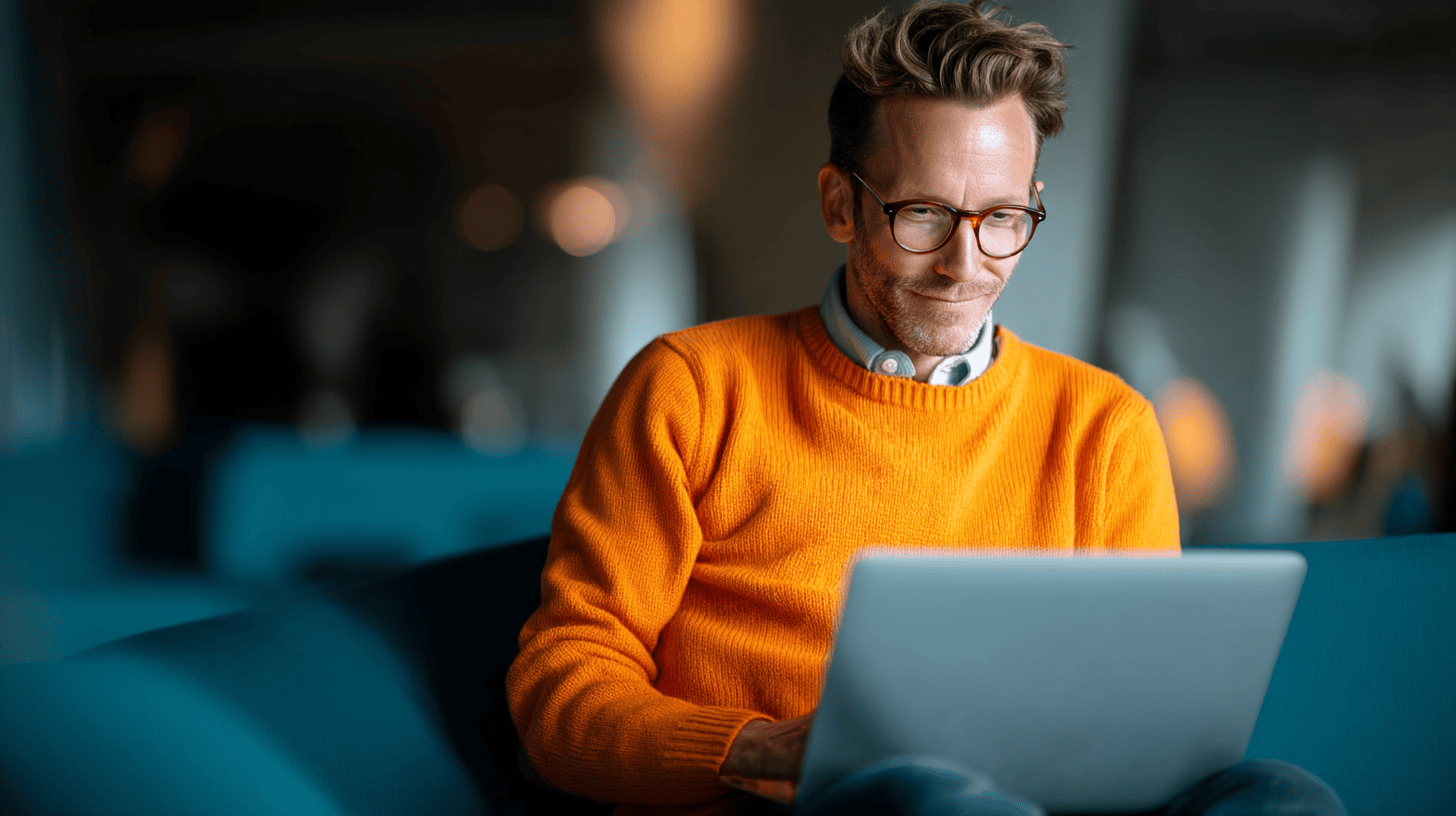 A man in an orange sweater uses a laptop while sitting on a teal couch in a modern, softly lit space.