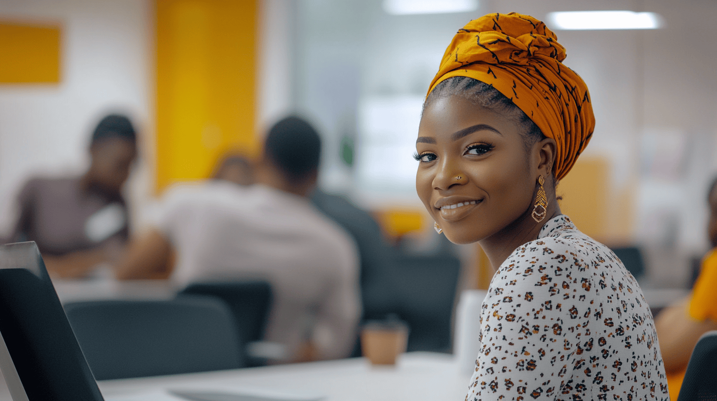 Smiling woman with an orange headwrap sits at a desk, with blurred people and bright colors in the background.