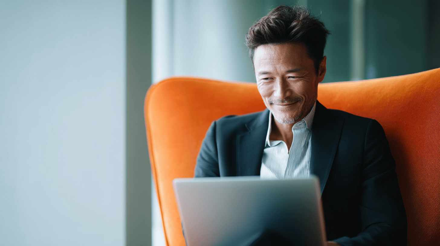 A man in a black suit sits in an orange chair, smiling while using a laptop in a bright, modern space.